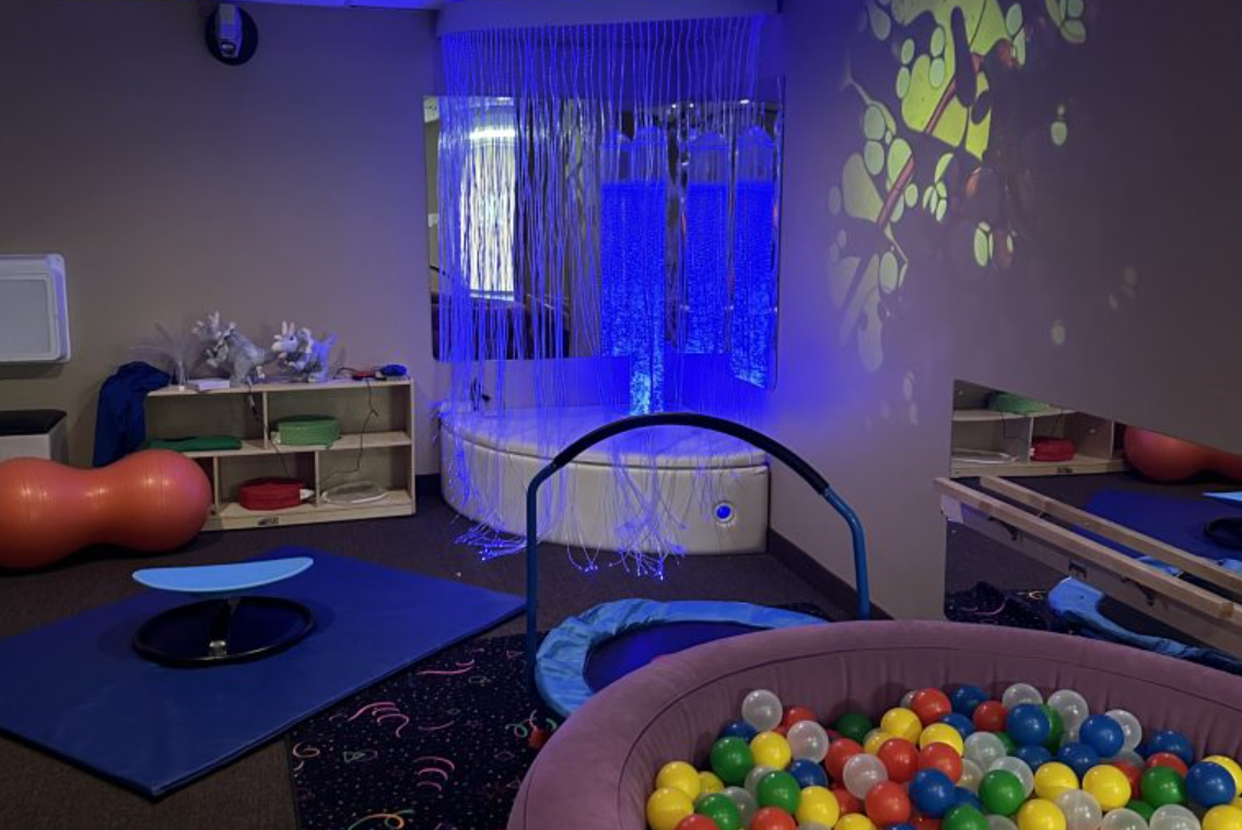 Image shows a corner of the sensory room, a ball pit in the foreground, a mini trampoline in the midground, and raised corner pad with a bubble tube coming out of it. Strands of florescent blue strips surround the pad.