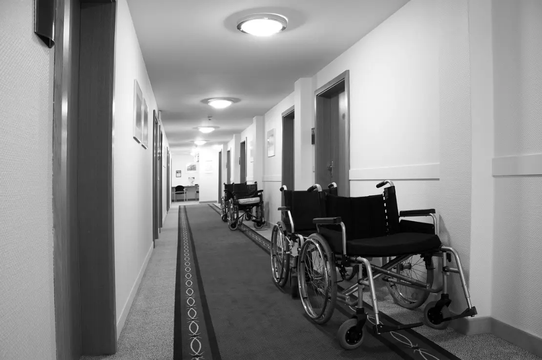 A stock photo is used adjacent to the NHTD article. It displays a long, narrow indoor hallway in a healthcare or assisted living facility, shown in black and white. The corridor has smooth, light-colored walls with several closed doors on both sides, each with a handle and small signage. Overhead, evenly spaced round ceiling lights illuminate the space. Along the right side of the hallway, multiple empty wheelchairs are lined up facing slightly toward the center, with metal frames, large rear wheels, and footrests visible. The floor is covered with a carpet that includes a darker central strip with a subtle patterned design running down the length of the corridor. At the far end of the hallway, a small desk or table and a chair are visible against the back wall, adding depth and perspective. The overall scene feels quiet, orderly, and institutional.