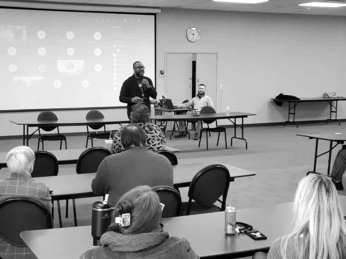Above this article, a photo is shown of the Reverend Doctor Damond A. Wilson, a Black man in his thirties with a beard and glasses wearing dark clothing, addressing STIC. He is standing in front of a projection screen showing a computer desktop and several tables and chairs. To Reverend Wilson’s right, a White man in a light top with a heavy beard operates a desktop beneath a clock reading 9:32. In the foreground we see the backs of the heads of audience members.