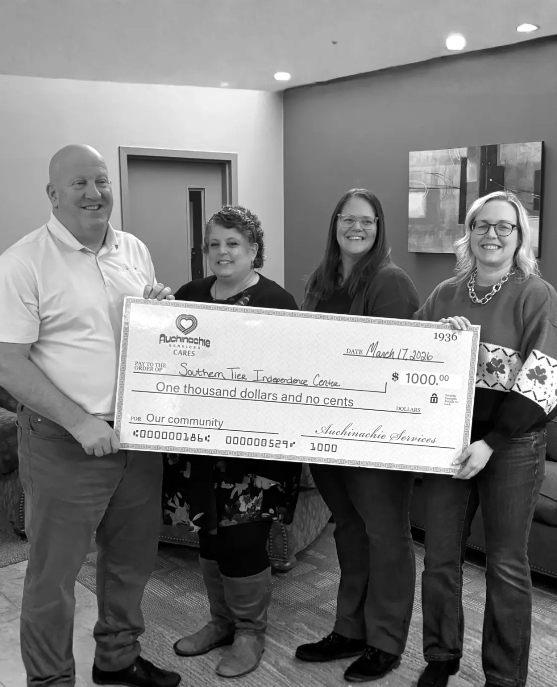A black-and-white photograph shows four adults standing indoors, smiling and holding a large ceremonial check together. The group is positioned in a hallway or office space, with a door visible behind them on the left and a framed abstract artwork on the wall to the right.